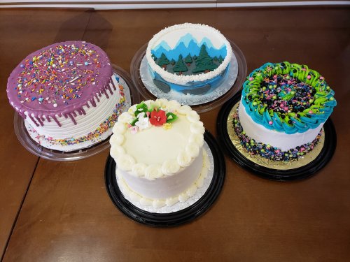 Five colorful cakes on a wooden table, featuring sprinkles, flowers, mountains, and vibrant frosting designs.