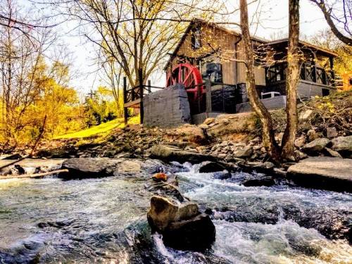A scenic view of a rustic building with a waterwheel beside a flowing river, surrounded by trees in autumn colors.