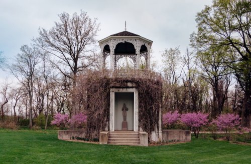 A gazebo surrounded by trees and pink flowers, with a statue at its entrance, set in a lush green landscape.