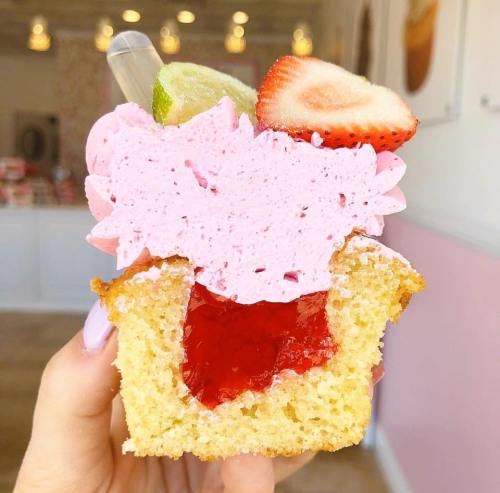 A hand holding a cupcake with pink frosting, strawberry, lime, and a jelly filling visible inside.