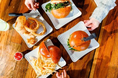 A table with two burgers, fries, onion rings, and hands reaching for the food.