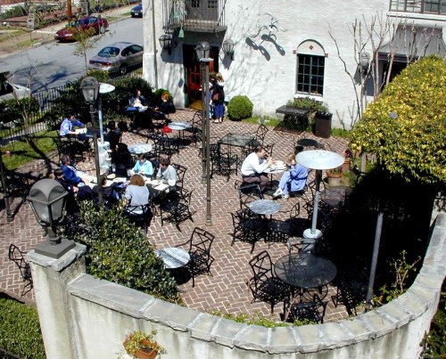 Outdoor café scene with people sitting at tables, surrounded by greenery and brick pathways.