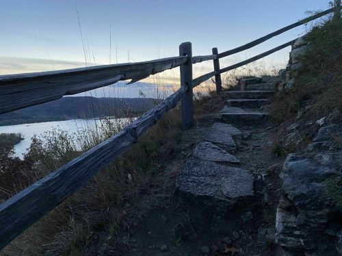 A winding stone path leads up a hillside, bordered by a wooden fence, with a serene view of the river at dusk.