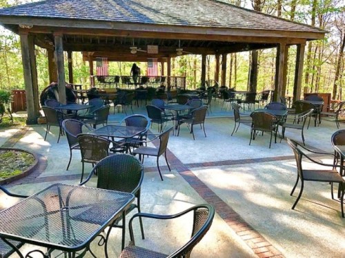 Outdoor seating area with black metal tables and chairs under a wooden pavilion surrounded by trees.