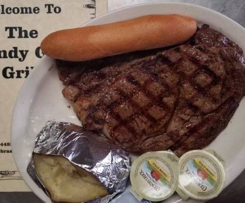 A plate with a grilled steak, a baked potato with foil, a bread roll, and two butter packets.