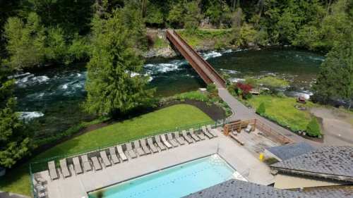 Aerial view of a pool area beside a river, with a wooden bridge and lush greenery surrounding the scene.