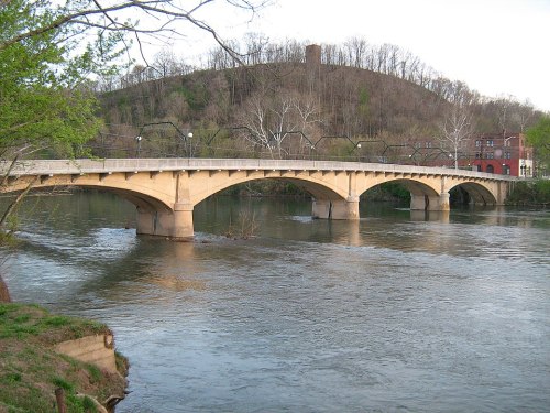 A stone bridge arches over a river, with trees and a hill in the background, showcasing a serene landscape.