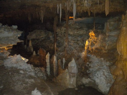 A dimly lit cave interior featuring stalactites and stalagmites, with rocky formations and a small pool of water.