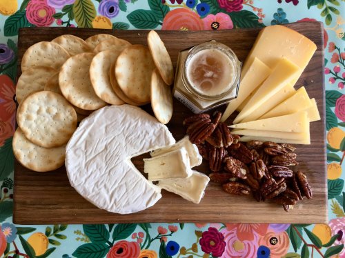A wooden board with assorted cheeses, crackers, pecans, and a jar of honey on a floral background.