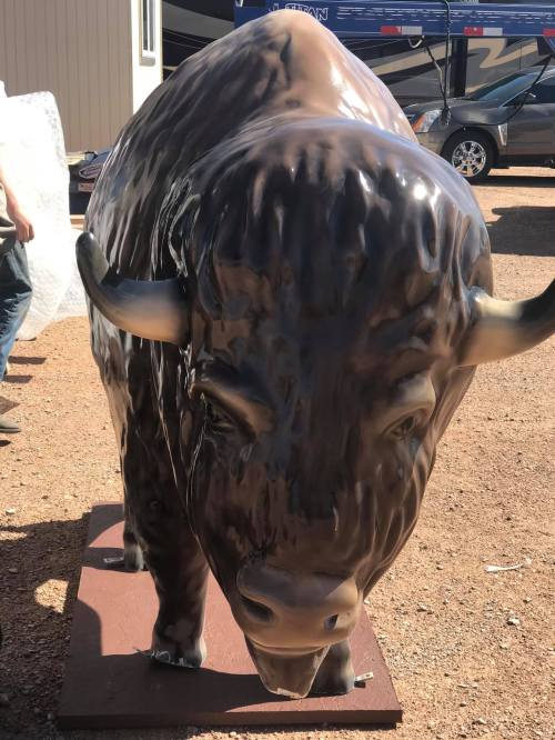 A large, realistic statue of a buffalo, positioned outdoors on a gravel surface.