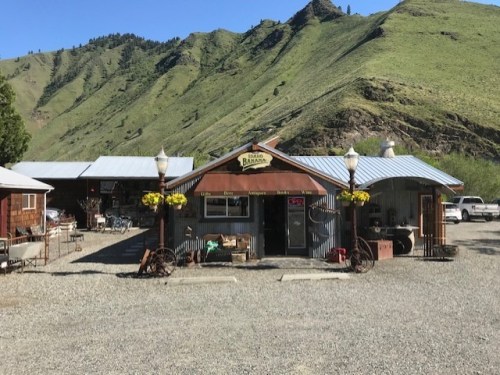 A rustic shop with a wooden facade, surrounded by green hills and gravel, featuring vintage items and flower baskets.