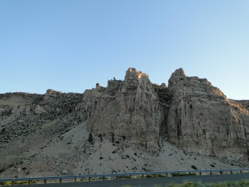 Rocky cliffs rise against a clear blue sky, with a highway guardrail in the foreground.