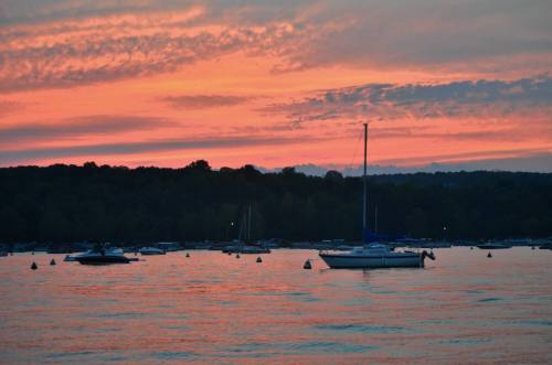 A serene sunset over a lake, with sailboats and moored vessels silhouetted against vibrant orange and pink skies.