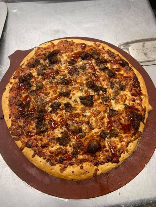 A freshly baked pizza topped with cheese, meatballs, and drizzled with barbecue sauce on a wooden pizza peel.