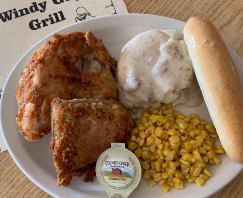 A plate of fried chicken, mashed potatoes with gravy, corn, a bread roll, and a butter packet on a wooden table.