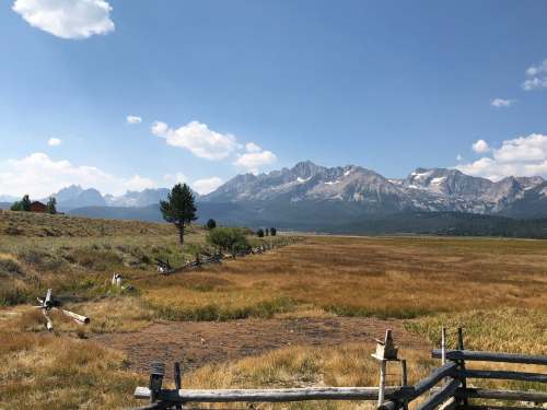 A scenic view of a grassy field with a wooden fence, mountains, and a clear blue sky in the background.