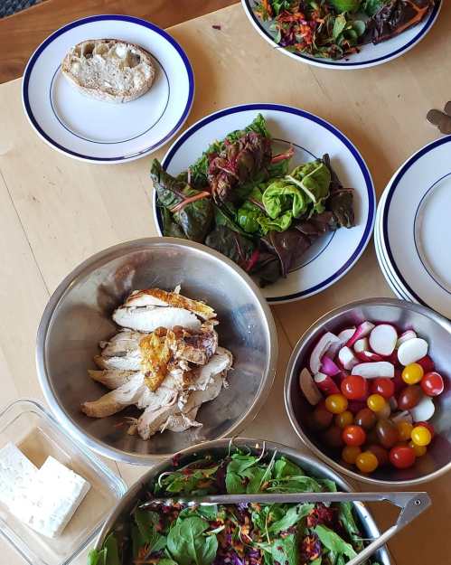 A table set with plates of salad, sliced chicken, cherry tomatoes, radishes, and a loaf of bread.