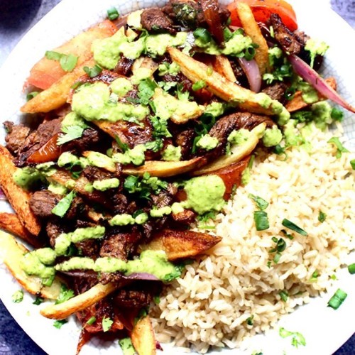 A plate of beef stir-fry with fries, topped with green sauce, served alongside brown rice and garnished with herbs.