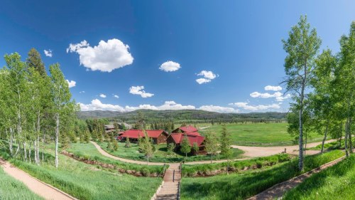 A panoramic view of a green landscape with a red-roofed building, trees, and a clear blue sky with fluffy clouds.