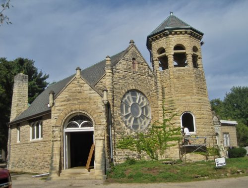 Historic stone church with a round window and bell tower, surrounded by trees and scaffolding for restoration.