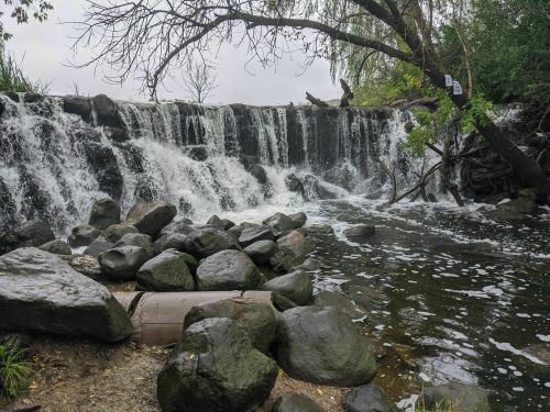 A waterfall cascades over rocks into a calm pool, surrounded by trees and greenery on a cloudy day.