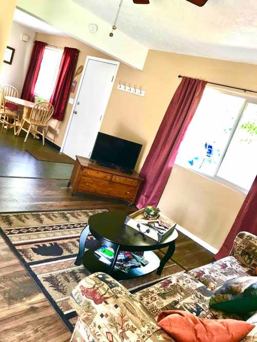 Cozy living room with a sofa, coffee table, TV, and dining area visible through an open doorway. Natural light from windows.