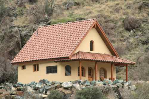 A small, rustic house with a tiled roof, set against a rocky hillside and surrounded by greenery.