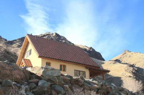 A cozy house with a tiled roof sits among rocky terrain, surrounded by mountains under a clear blue sky.