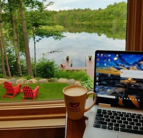 A cozy workspace by a lake, featuring a laptop, a coffee mug, and red chairs on the shore surrounded by greenery.