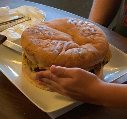 A large burger on a plate, with a hand reaching towards it, showcasing its size and toppings.