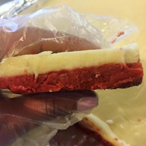A hand holding a slice of layered red and white dessert, partially wrapped in plastic.