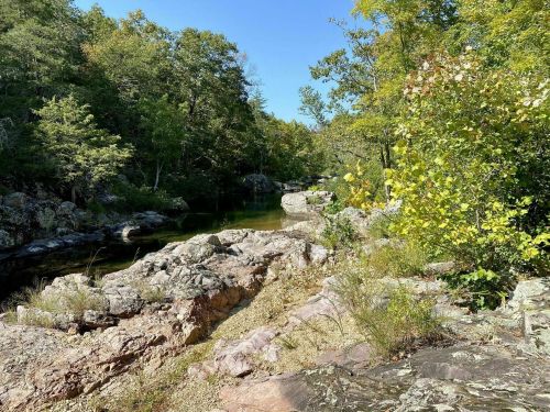 A serene landscape featuring a calm water body surrounded by rocky terrain and lush green trees under a clear blue sky.