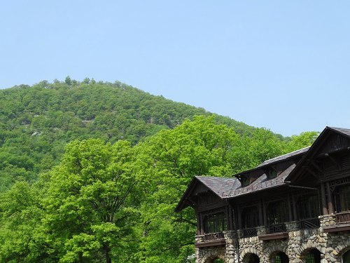 A stone building with a wooden roof surrounded by lush green trees and a mountain in the background under a clear sky.