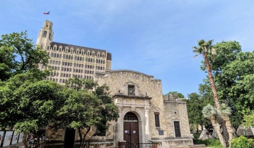 Historic stone building with a decorative entrance, surrounded by trees, with a tall building and palm trees in the background.