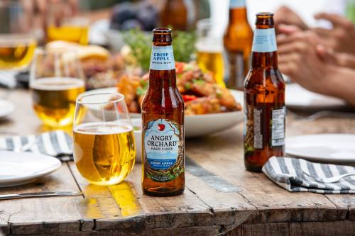 A table set with food and drinks, featuring a bottle of Angry Orchard hard cider and glasses of beer.