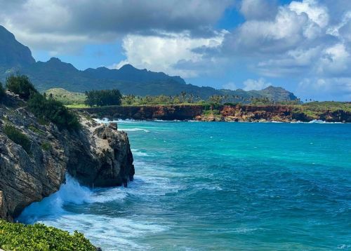 A scenic coastal view with rocky cliffs, turquoise waters, and lush green mountains under a partly cloudy sky.