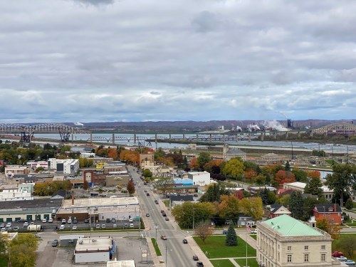 A scenic view of a river with bridges, surrounded by autumn foliage and a cityscape under a cloudy sky.