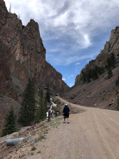 A person walks along a dirt road between towering rocky cliffs and trees, with a stream visible nearby.