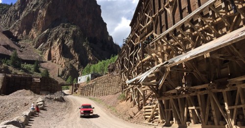 A red truck drives along a dirt road beside wooden structures in a mountainous landscape.