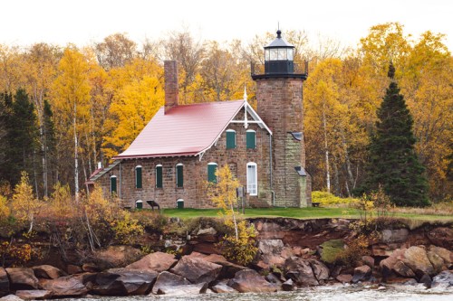 A stone lighthouse with a red roof, surrounded by autumn foliage and rocky shoreline.