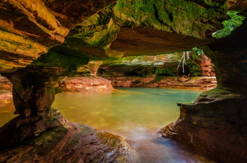A serene natural cave with smooth rock formations and a tranquil pool of water reflecting greenery.