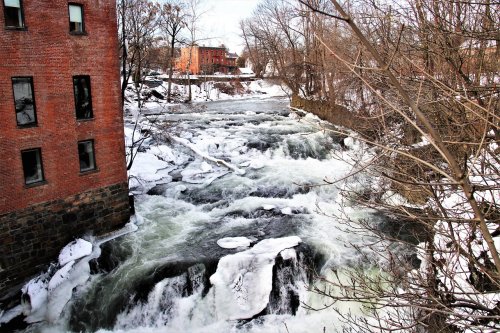 A rushing river with ice and snow, bordered by a brick building and bare trees in a winter landscape.