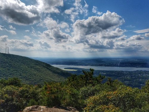 A scenic view of a lush green mountain overlooking a river, with clouds scattered across a blue sky.