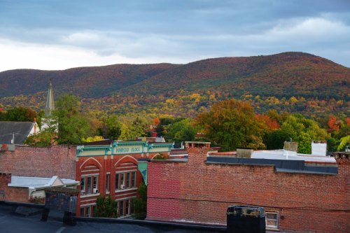 A view of colorful autumn trees on mountains, overlooking brick buildings in a small town under a cloudy sky.