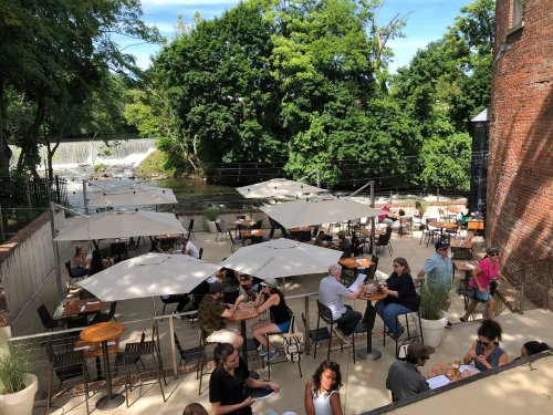 Outdoor dining area with umbrellas, people enjoying meals, and a scenic view of a river and greenery in the background.