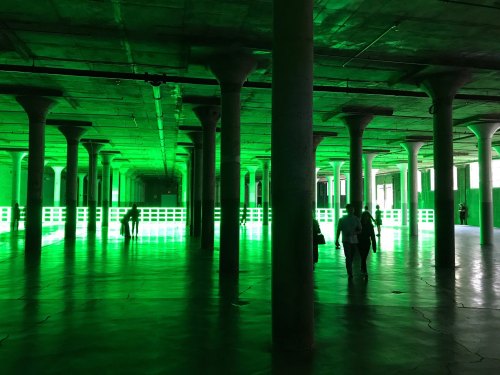 A spacious industrial room with green lighting, featuring tall columns and people walking through the illuminated space.