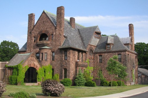 A large, historic stone mansion with ivy-covered walls and multiple chimneys, set against a clear blue sky.