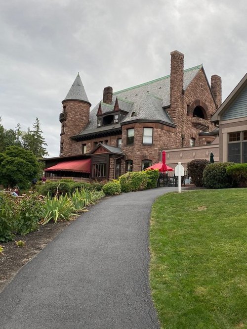 A large stone mansion with a turret, surrounded by greenery and a winding path leading to its entrance.