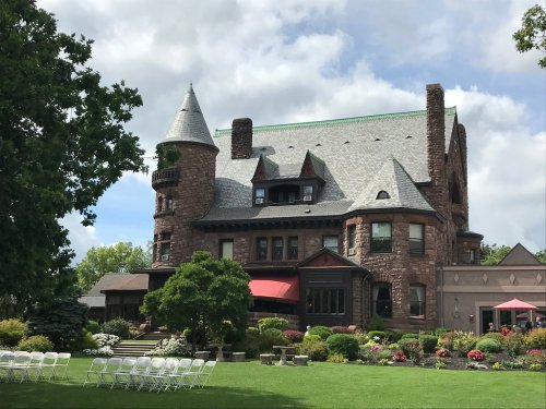 A large, historic stone mansion with a turret, surrounded by lush greenery and colorful flower beds.