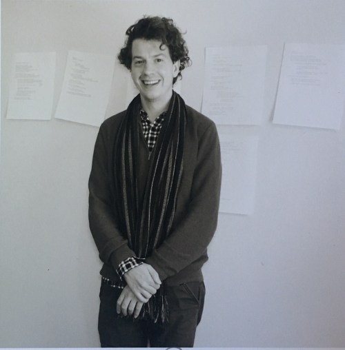 A smiling person with curly hair stands in front of papers on a wall, wearing a scarf and a sweater.